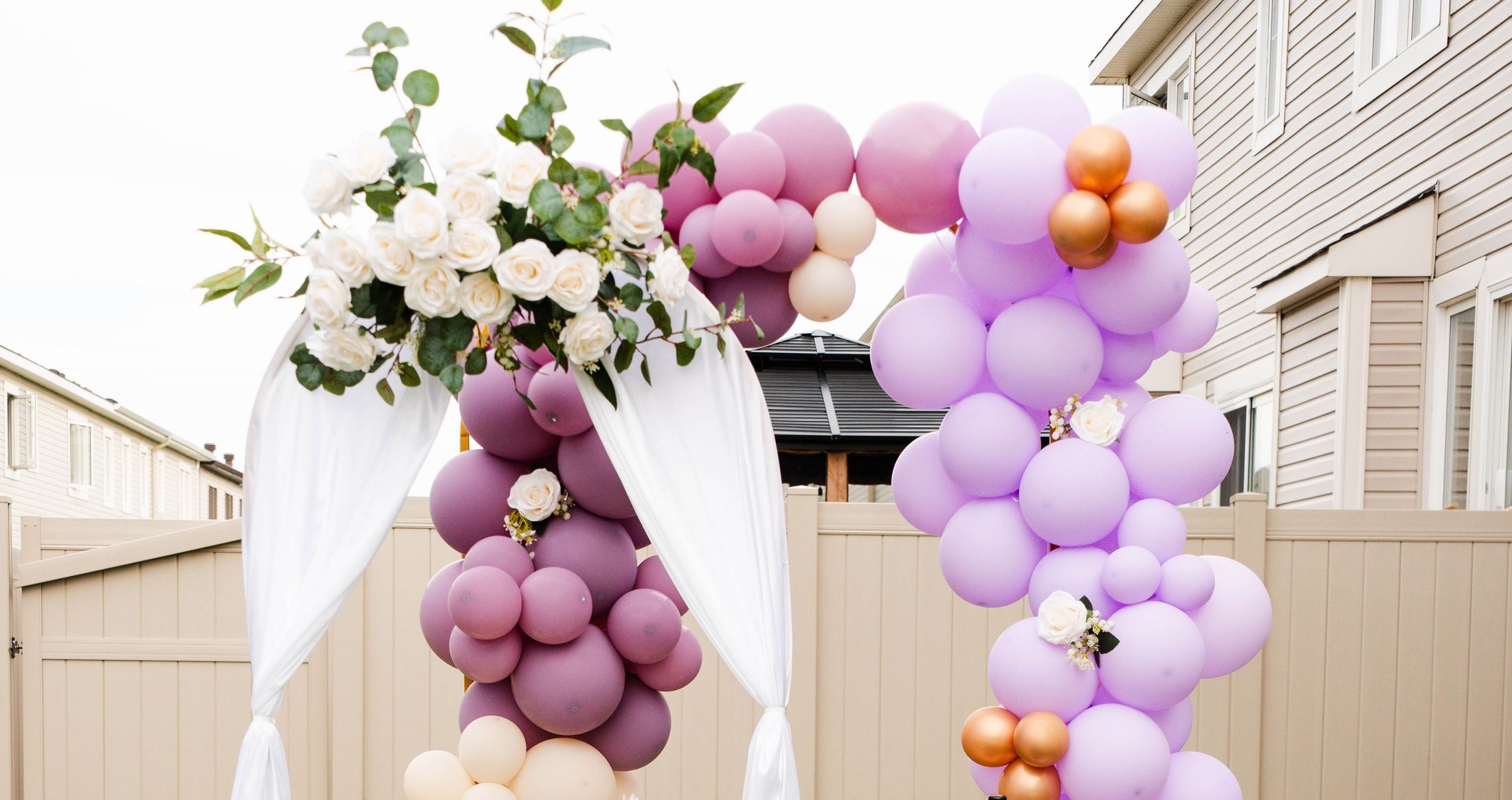 Lilac, mauve and cream balloon arch with white roses and draped fabric in a backyard