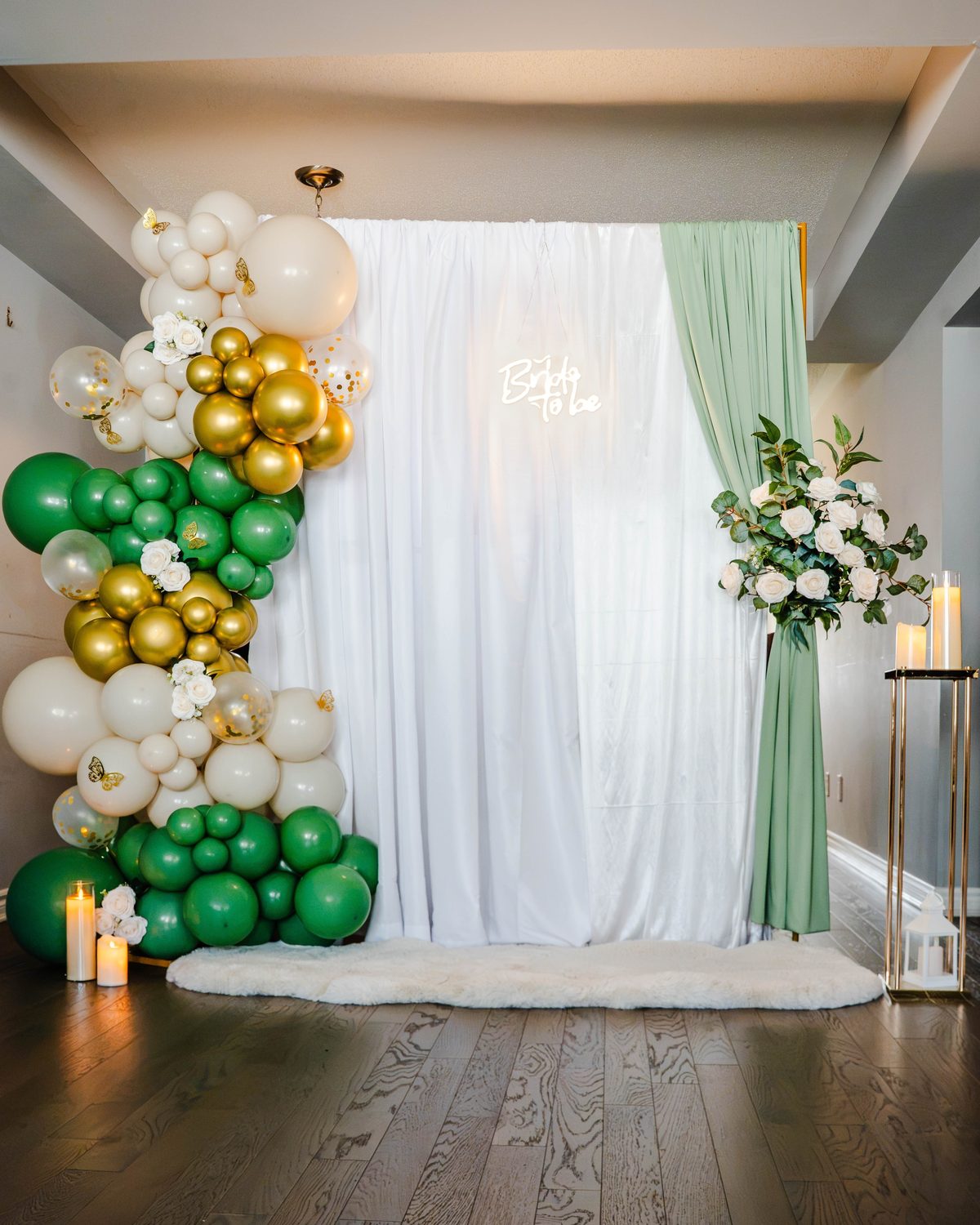 Green, gold and cream balloon garland beside a white drape with a glowing Bride to be neon sign and a sage curtain with white roses