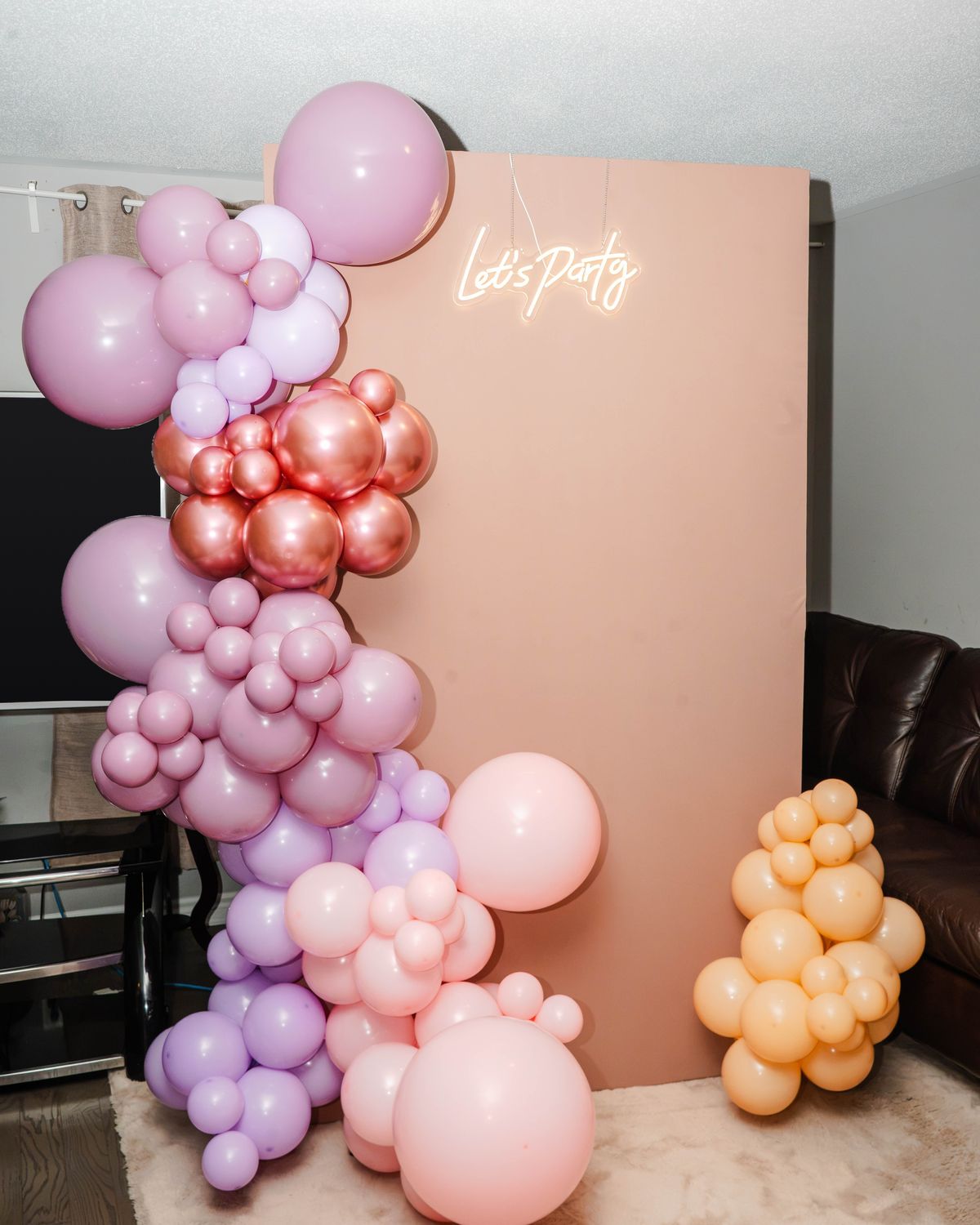 Balloon garland in mauve, lilac, blush and rose gold framing a peach backdrop with a Let's Party neon sign